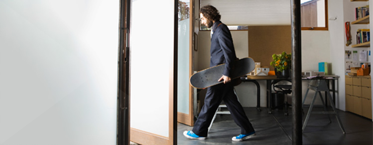 man with long hair, beard, suit, and sneakers walking outside with skateboard 