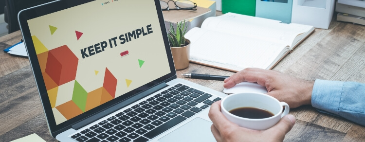 Keep It Simple on laptop screen. desk shows plant, glasses, mouse, book, pen, and hand holding coffee mug