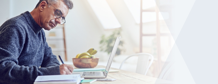 man writing with pen on notepad while working on laptop in sunny room